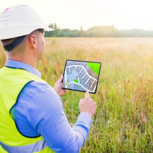 A builder stands in an empty field holding a tablet displaying plans for a new housing development.