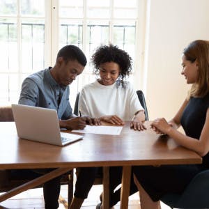 A real estate agent sits at a table with a Black couple signing a contract.