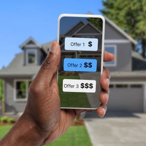 A person holds a smartphone displaying three offers on a house, with a home in the background