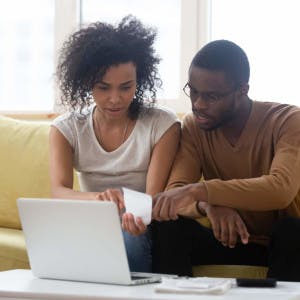 A Black couple sits on their couch and looks at homes on a laptop.