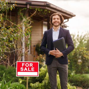 A young male real estate agent stands smiling in front of a home for sale.