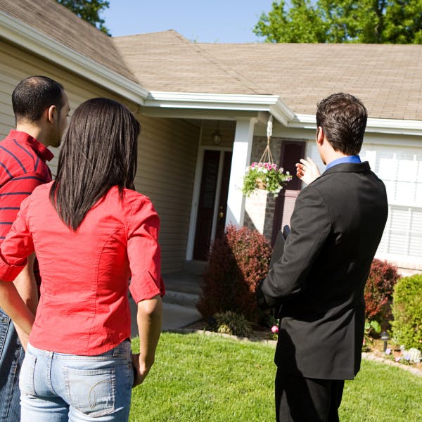 A couple and a real estate agent stand in a yard looking at the front of a house. 
